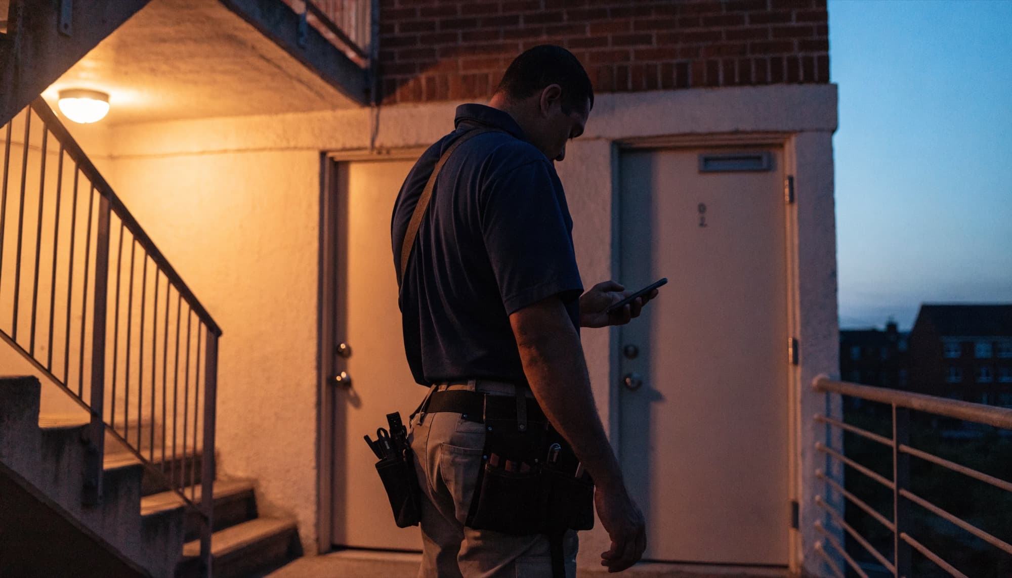 A maintenance technician checking his phone at a unit door at dusk.