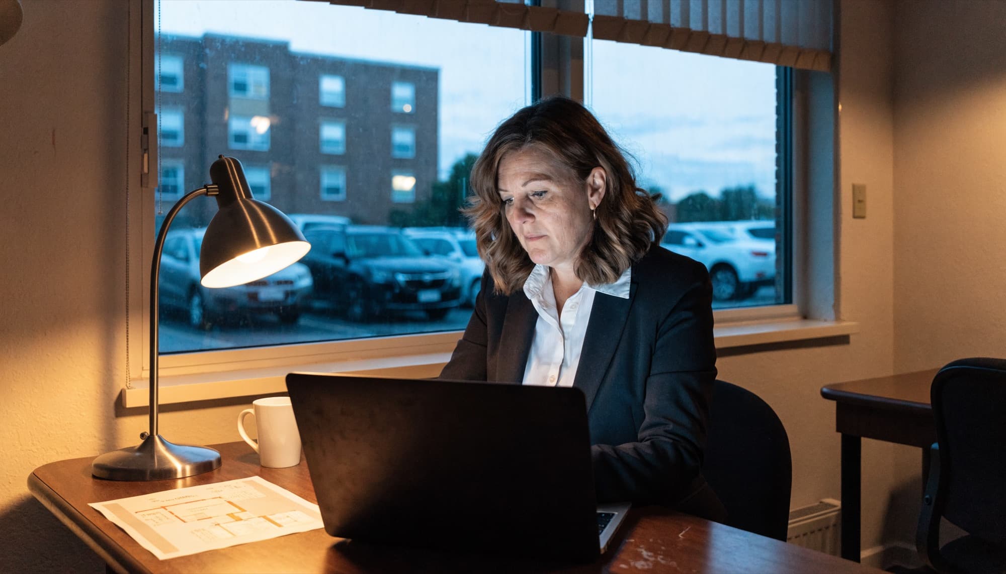 A property manager working at a laptop by the office window in the evening.