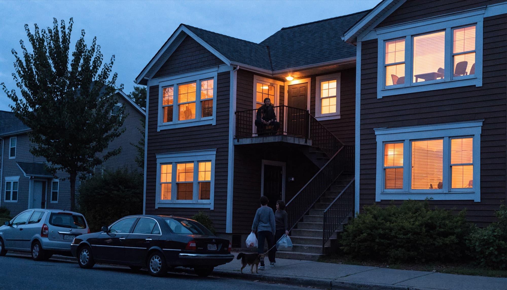 Residents coming home at dusk to a two storey building with lit windows.