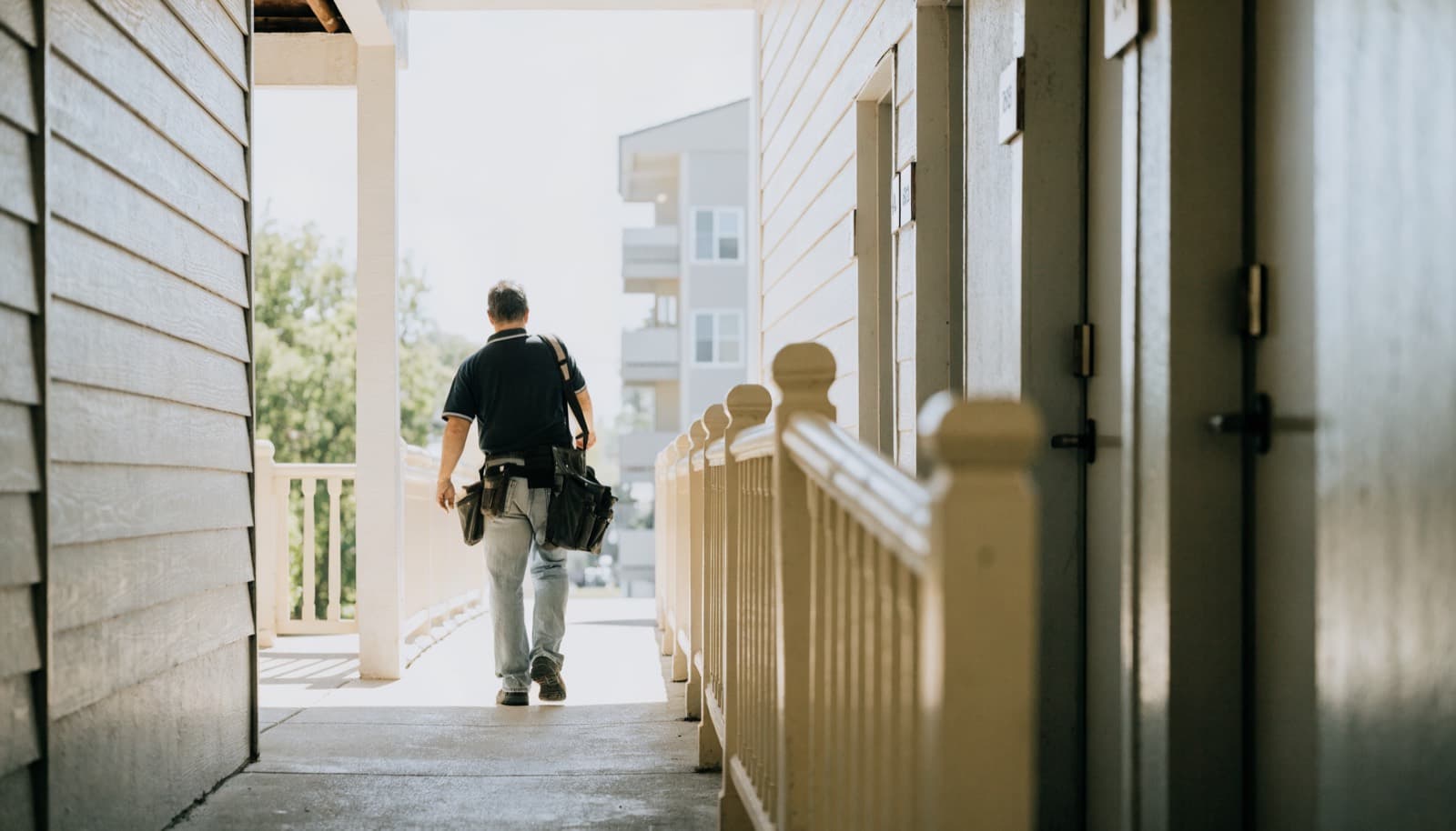 A maintenance technician walking an exterior breezeway in daylight, tool bag in hand.