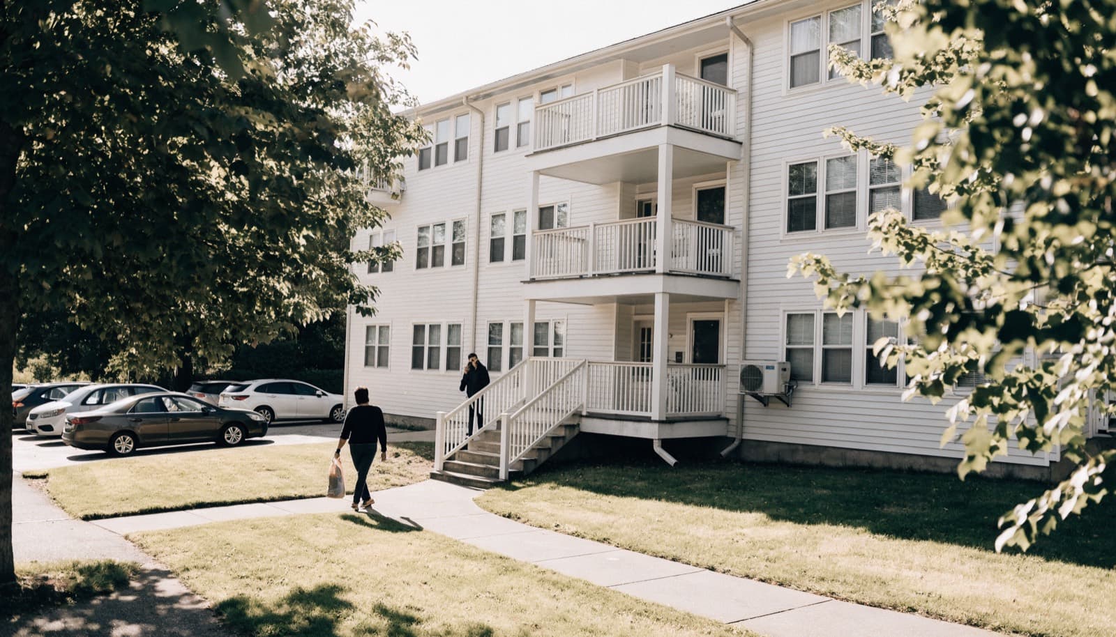 Residents outside a three storey apartment building on a bright afternoon, one walking the path, one on the phone by the stairwell.